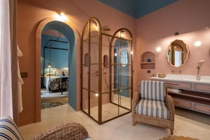 A room with a glass-enclosed shower, a wicker chair, a vanity with a mirror, and a bed in the background. - Palazzo Ceraselli (Martina Franca)