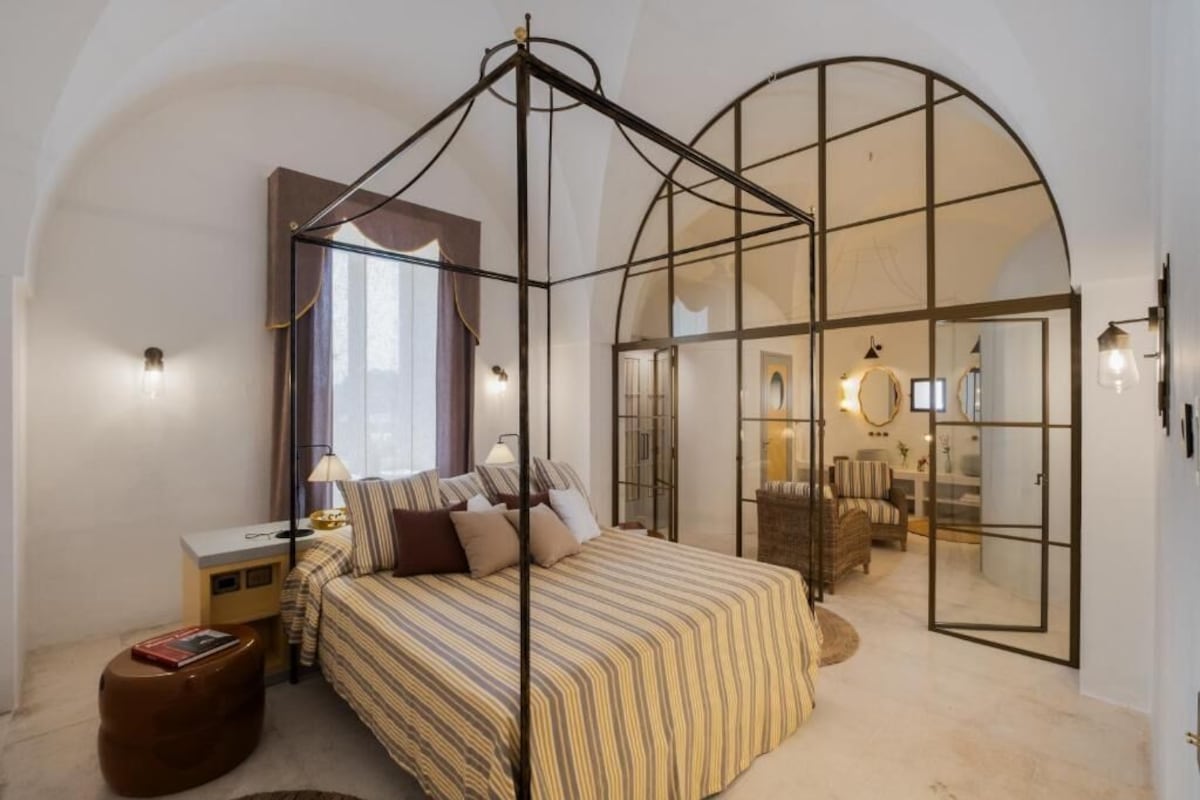 A bedroom with a canopy bed, a small bedside table, a reading chair, and a large arched window.