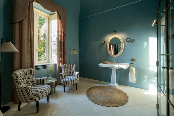 A room with a striped armchair, a round rug, a white pedestal sink, and a window with curtains.