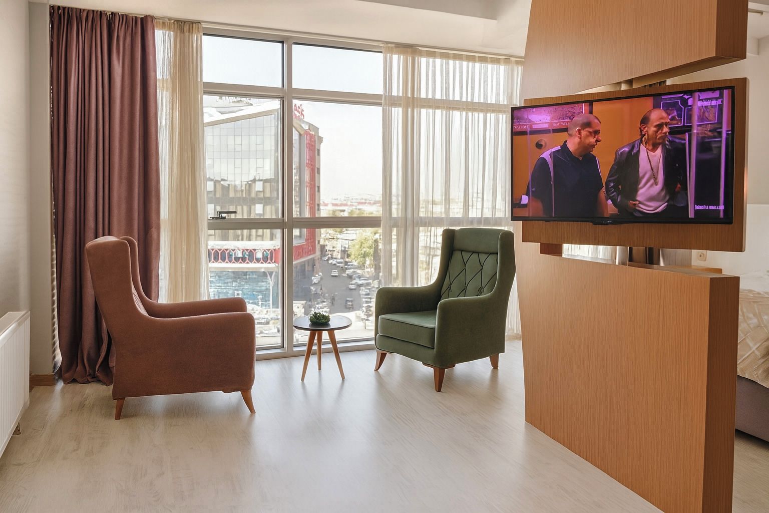 A modern hotel room with a large window, two armchairs, a small table, and a flat-screen TV.