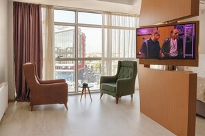A modern hotel room with a large window, two armchairs, a small table, and a flat-screen TV.