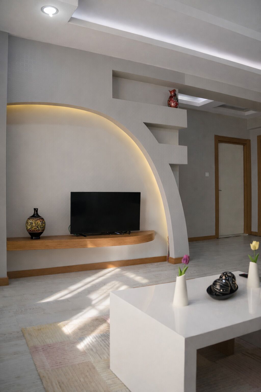 A modern living room with a flat-screen TV on a built-in shelf, a vase with flowers, and a decorative bowl on a white console table.