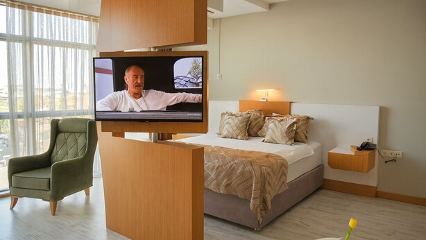A modern hotel room with a large bed, a television mounted on a wooden panel, a green upholstered armchair, and a bedside table with a lamp.