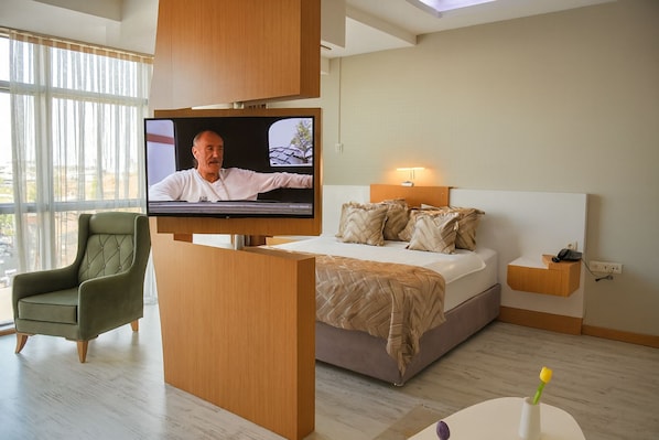 A modern hotel room with a large bed, a television mounted on a wooden panel, a green upholstered armchair, and a bedside table with a lamp. - THE CITY WALLS SUIT OTEL (Diyarbakir)