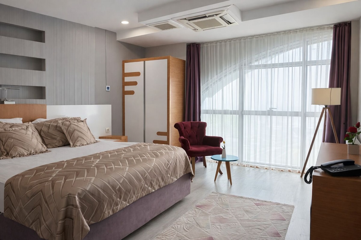 Una habitación de hotel moderna con una cama grande, un sillón rojo, una mesita y un escritorio con lámpara y teléfono.