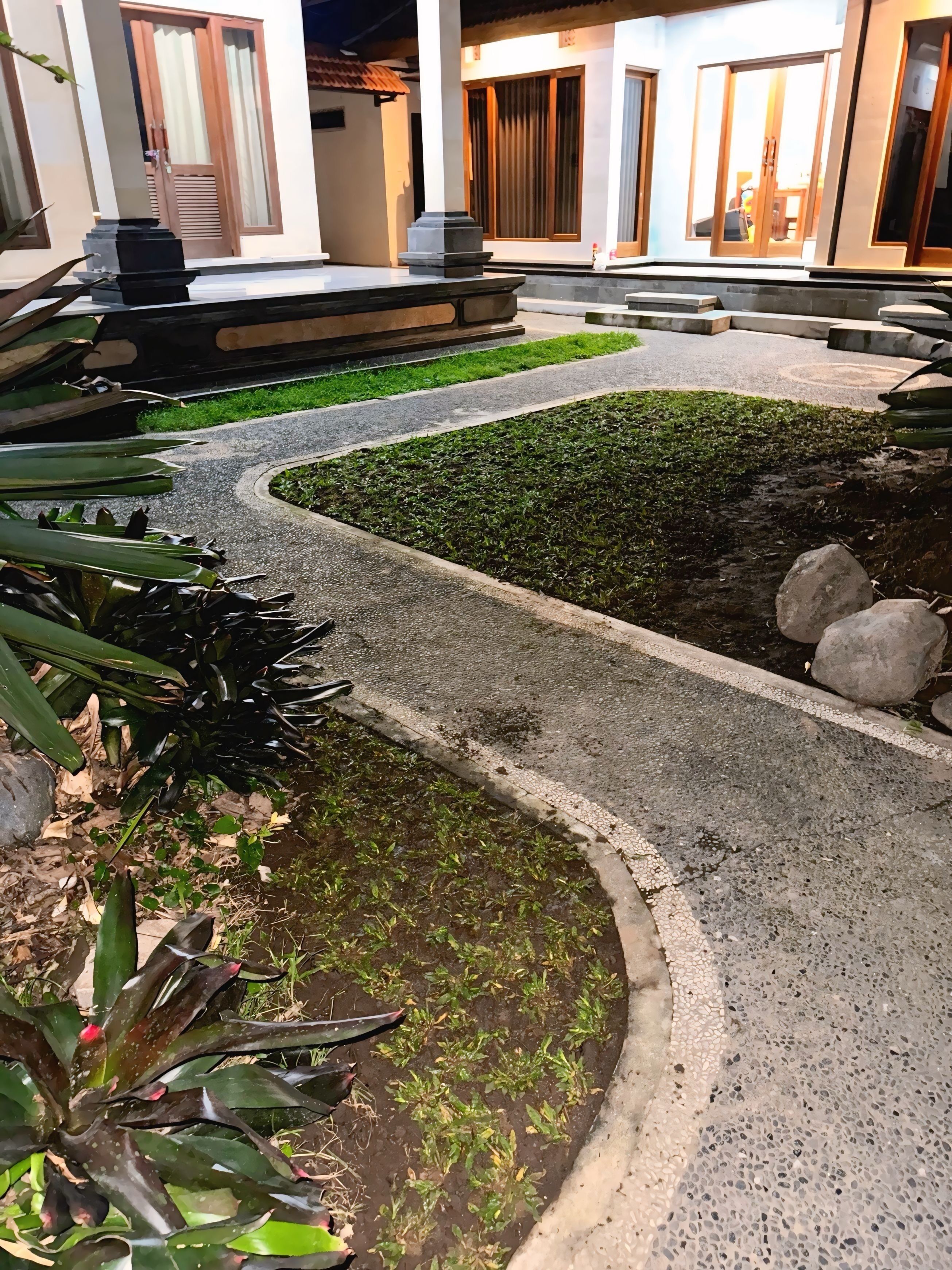 A pathway leading to a building entrance with a modern design, surrounded by greenery and a small garden area.