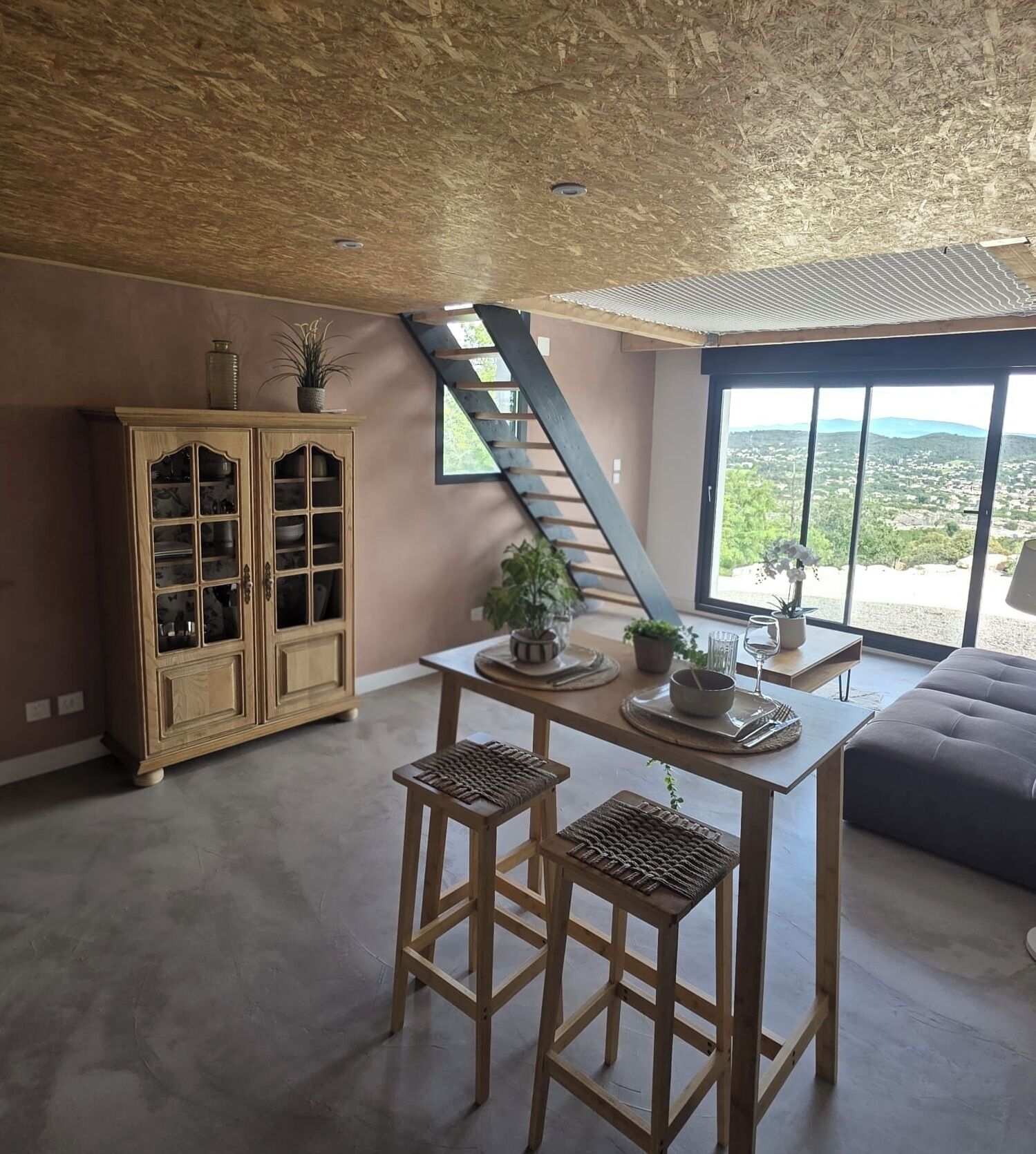 Ein Zimmer mit hoher Decke, einem Holzschrank, einem Tisch mit Hocker und Blick durch große Fenster.
