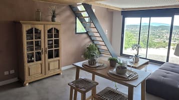 Ein Zimmer mit hoher Decke, einem Holzschrank, einem Tisch mit Hocker und Blick durch große Fenster.