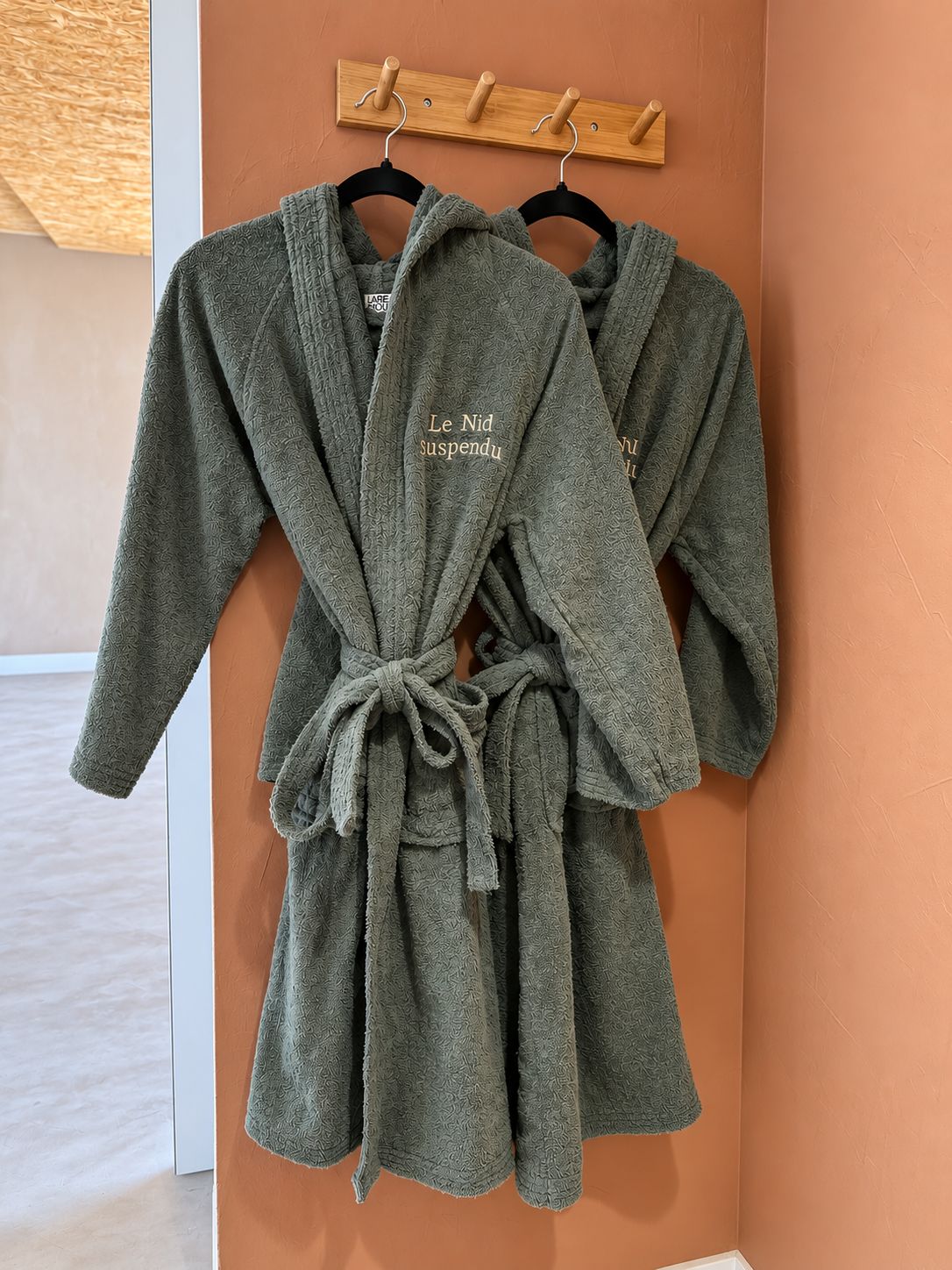 Two grey robes with a tied knot and a tag on one, hanging on a wooden rack against a peach-colored wall.