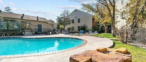 A swimming pool with a stone feature in the foreground, surrounded by a grassy area and a building with a sloped roof.