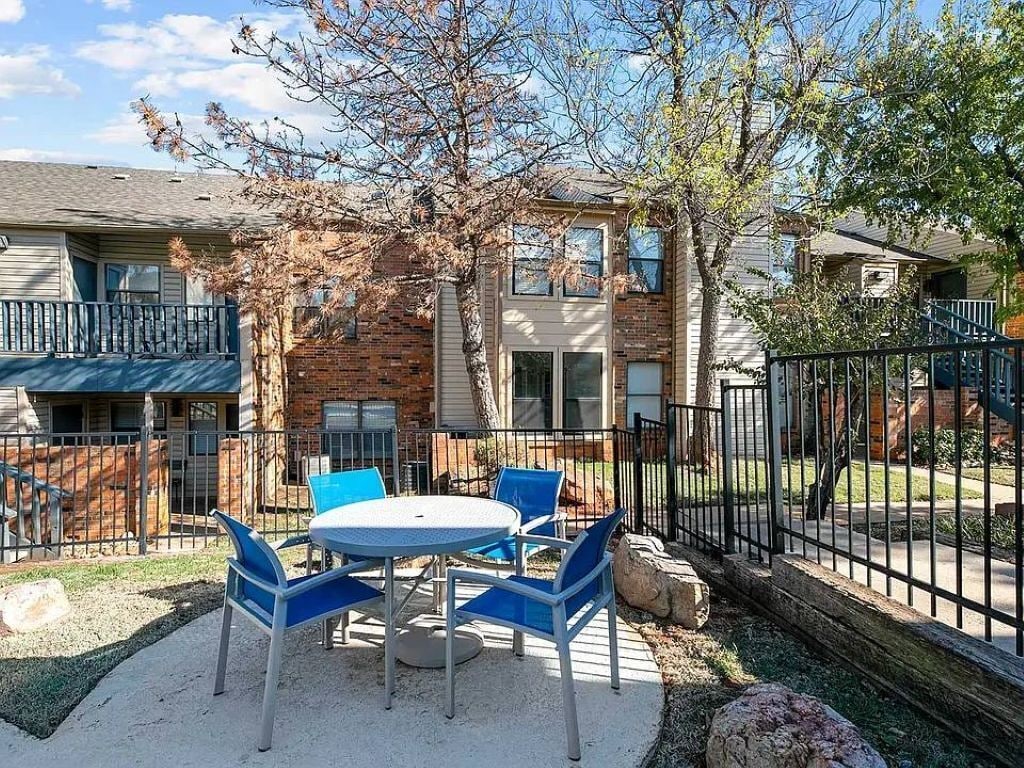 An outdoor patio with a round table and blue chairs, surrounded by a brick building with a balcony.