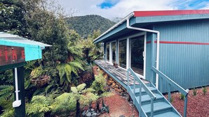 A building with a wooden deck and a staircase leading to a covered area, surrounded by lush greenery and a mountainous backdrop.