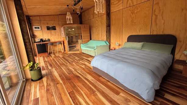 A modern bedroom with a wooden floor, a bed with green bedding, a desk with stools, and a kitchenette.