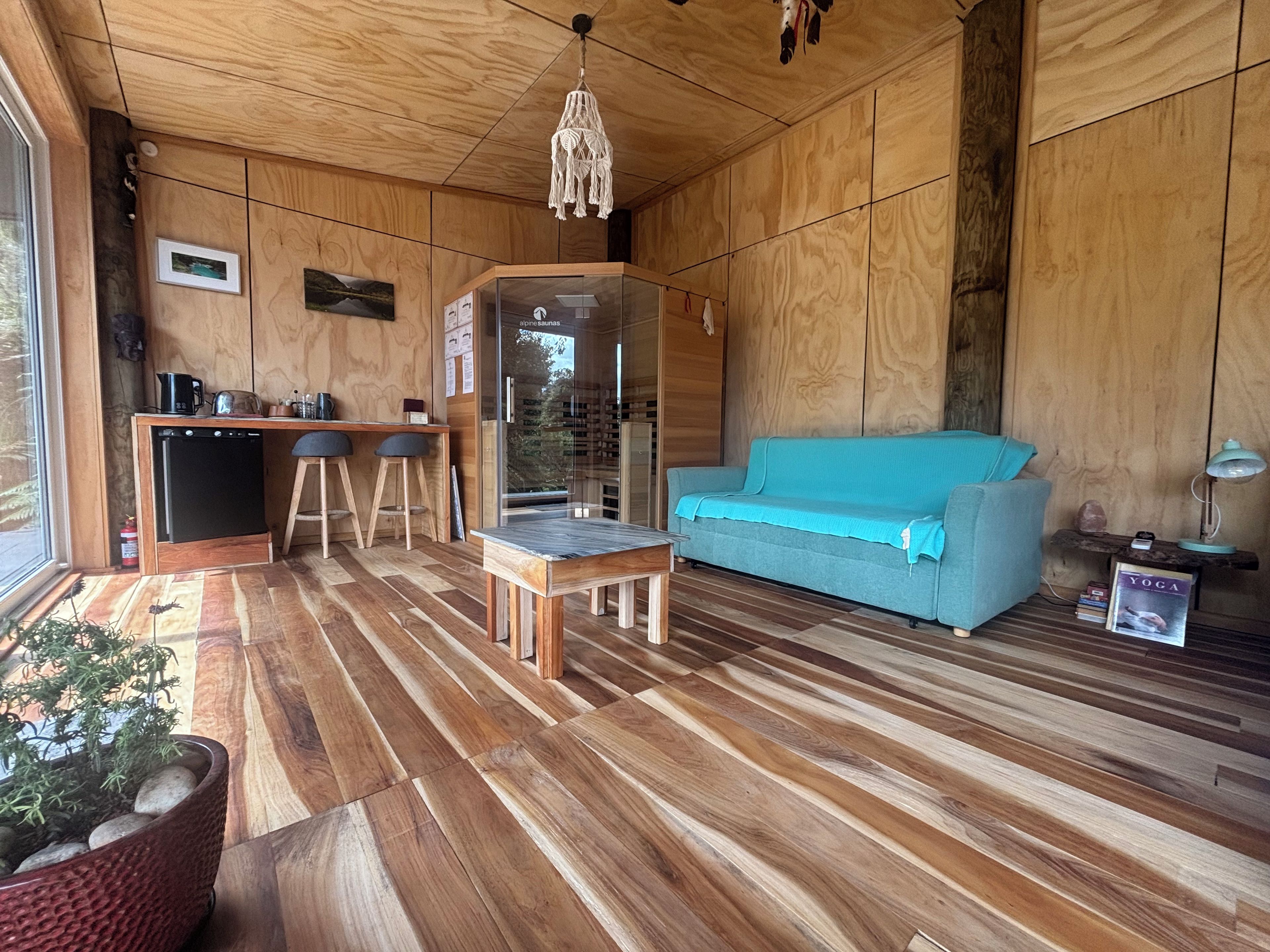 A wooden interior with a sofa, a small table, a kitchenette, and a view through a glass door.