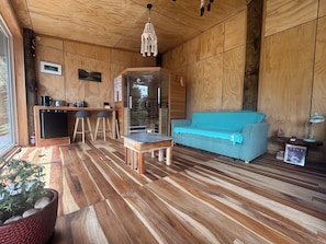 A wooden interior with a sofa, a small table, a kitchenette, and a view through a glass door.