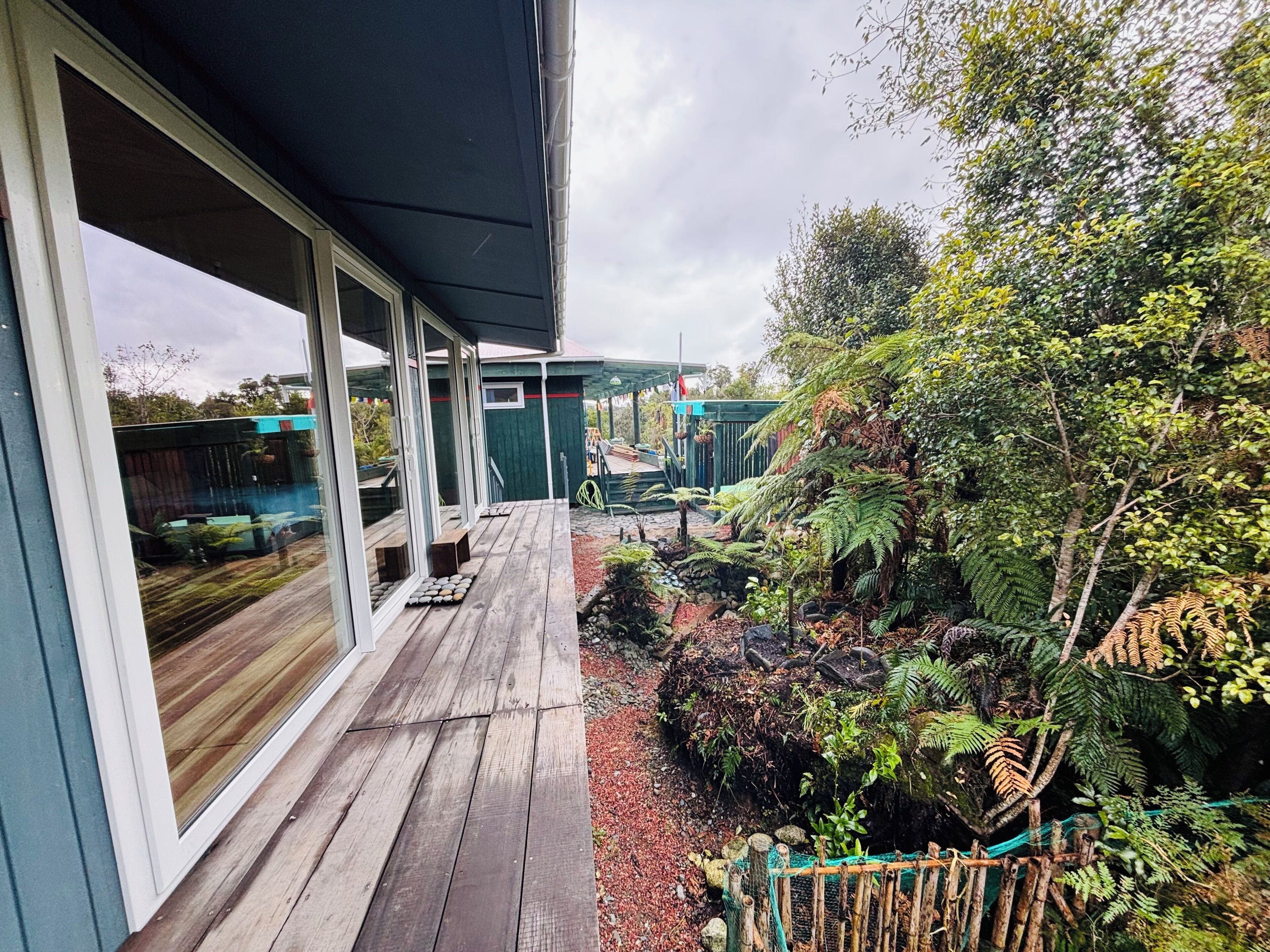 Eine Holzterrasse mit Glastüren, die zu einem Gartenbereich mit verschiedenen Pflanzen und einem grünen Zaun führen.