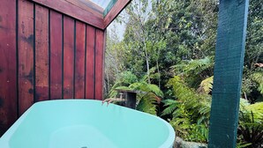 A large freestanding bathtub in a natural setting with a wooden structure and lush greenery in the background.