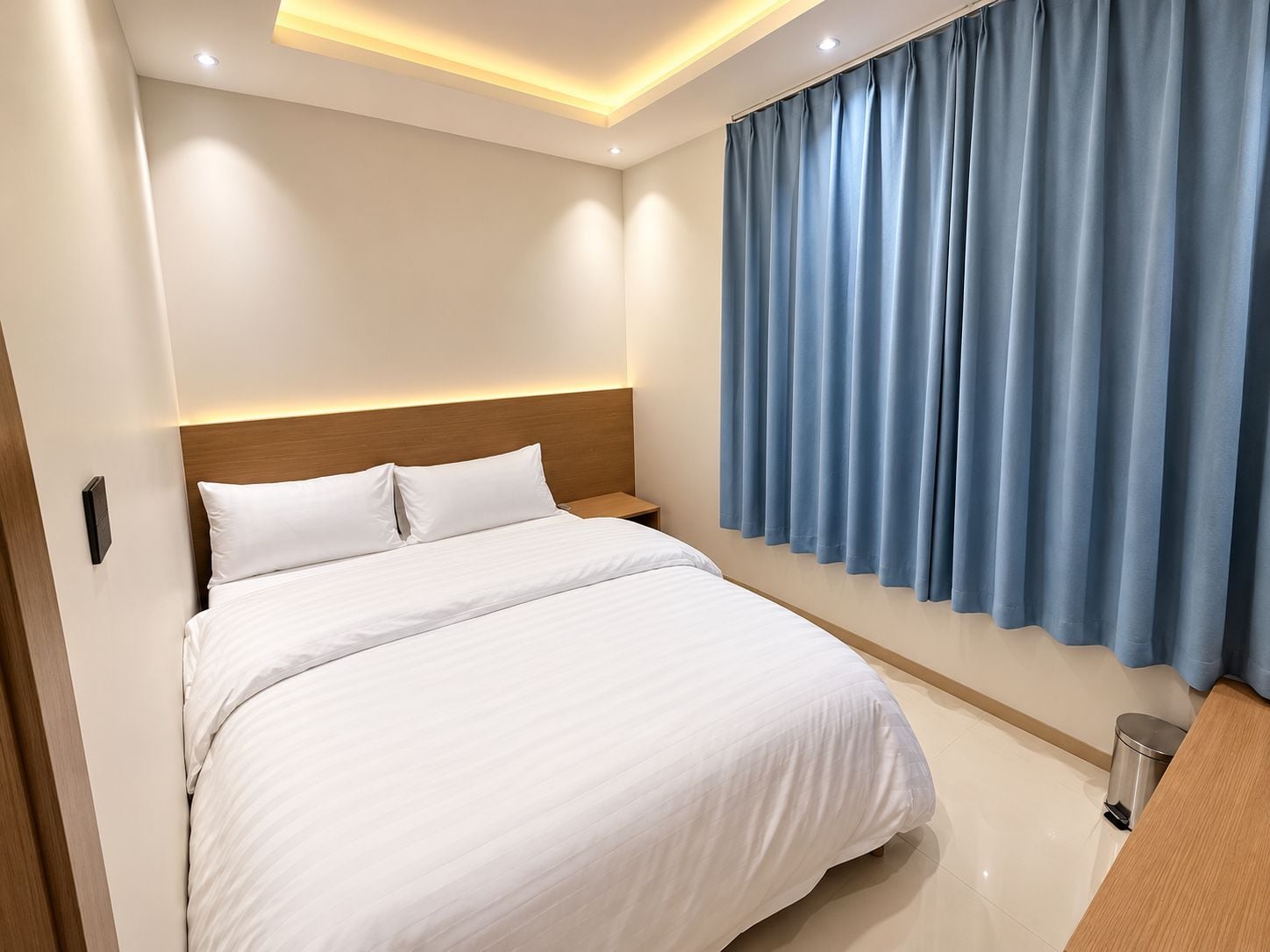 A hotel room with a bed, a nightstand, a trash bin, and a window with curtains.