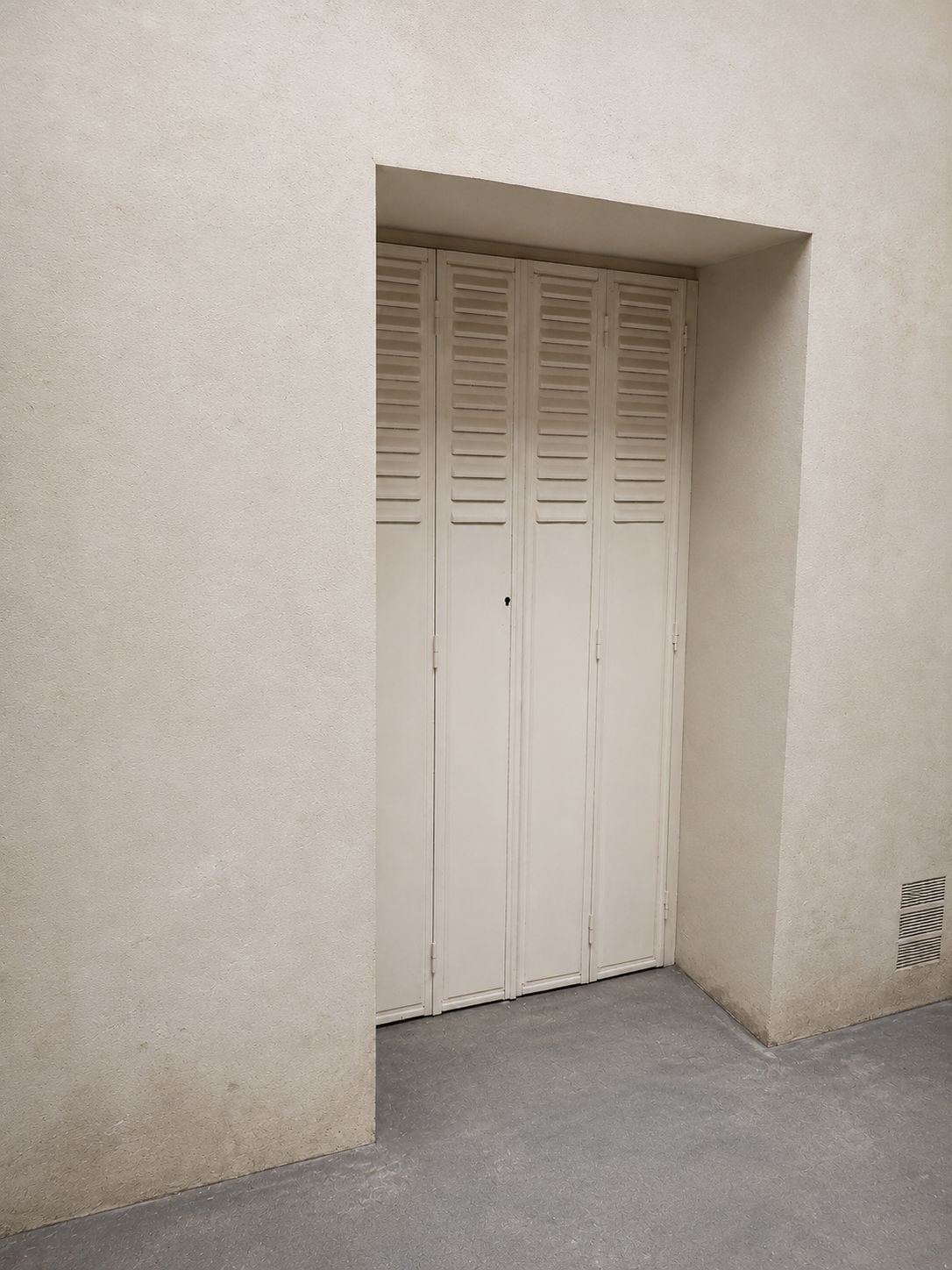 A closed door with horizontal louvered panels, a concrete floor, and a plain wall.