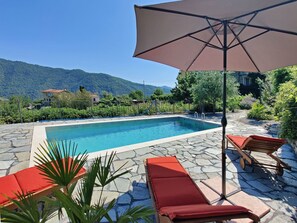 Uma piscina com espreguiçadeiras e um guarda-sol, rodeada por vegetação e montanhas.