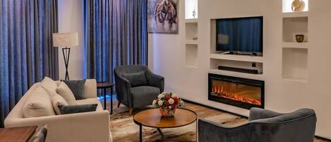A modern living room with a sofa, armchairs, a coffee table, a fireplace, a TV, and a painting on the wall.