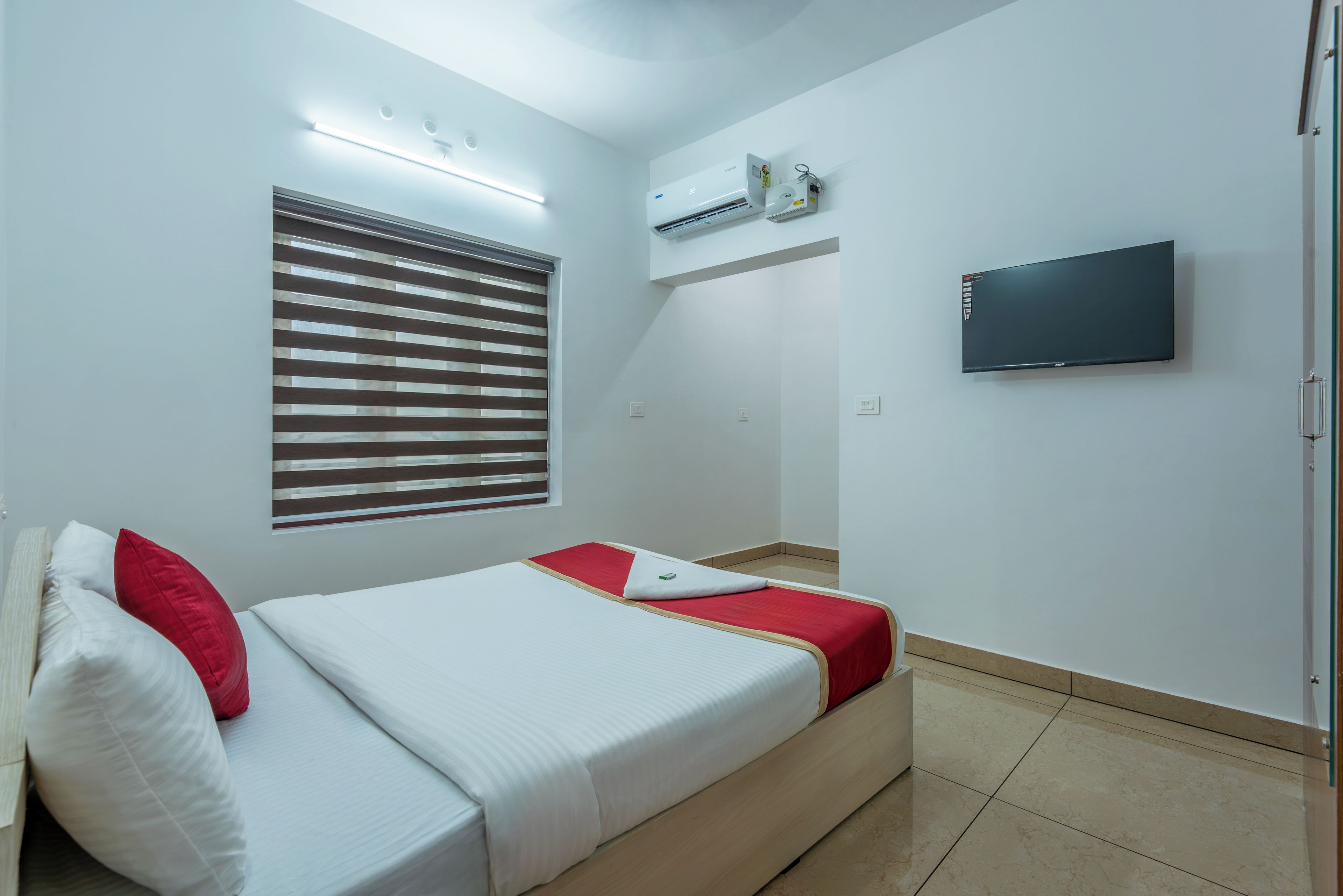 A hotel room with a bed, a TV, an air conditioning unit, and a window with blinds.