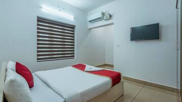 A hotel room with a bed, a TV, an air conditioning unit, and a window with blinds.