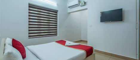 A hotel room with a bed, a TV, an air conditioning unit, and a window with blinds.