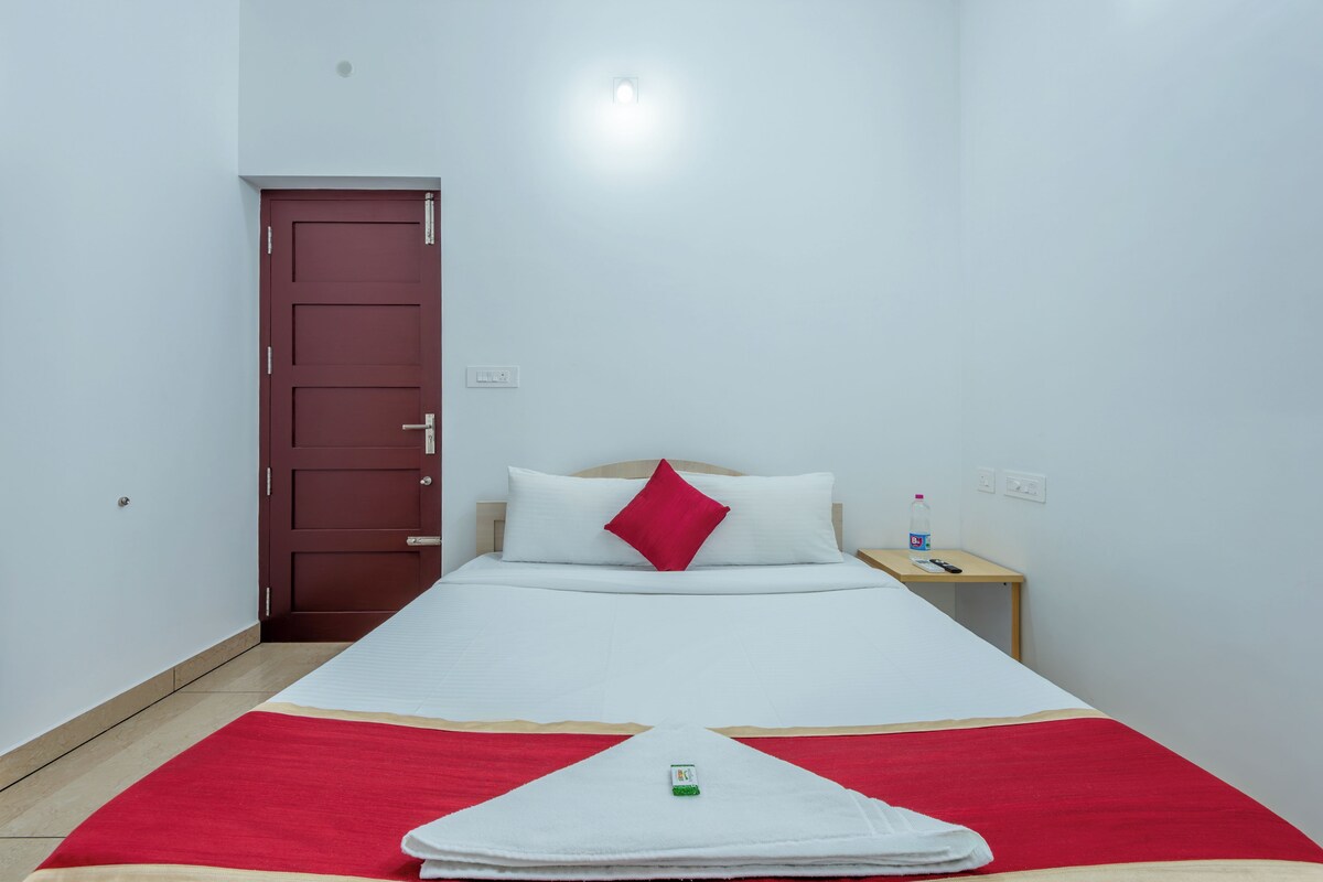 Habitación individual con una cama de madera, un edredón blanco y rojo, una mesita de noche con una botella y un control remoto, y una puerta roja.