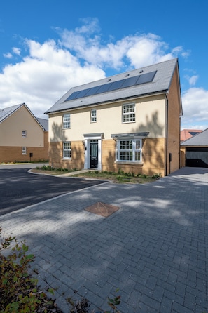 A two-story residential building with a brick facade, a flat roof with solar panels, and a paved driveway. - Five separate bedrooms on a quiet cul-de-sac in NR4, minutes from the A47. A room each for a team of five, or sleeps up to 9 when couples share. Open-plan kitchen the full depth of the house, driveway parking for three and 293 Mbps WiFi. (Norwich)