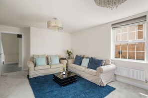 A living room with a beige sofa set, a blue rug, a chandelier, and a window with a view of a brick building. - Five separate bedrooms on a quiet cul-de-sac in NR4, minutes from the A47. A room each for a team of five, or sleeps up to 9 when couples share. Open-plan kitchen the full depth of the house, driveway parking for three and 293 Mbps WiFi. (Norwich)