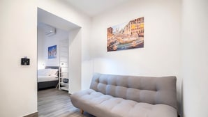 A modern hotel room with a grey sofa, a bed with white bedding, a painting of a cityscape, and a wooden floor.