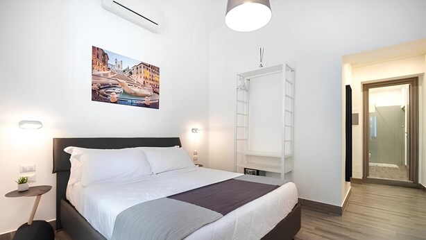 A modern hotel room with a large bed, bedside tables, a wall-mounted air conditioning unit, a ceiling light fixture, and a framed picture on the wall.
