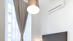 A modern bedroom with a bed, a hanging light fixture, an air conditioning unit, and a view through a window with curtains.