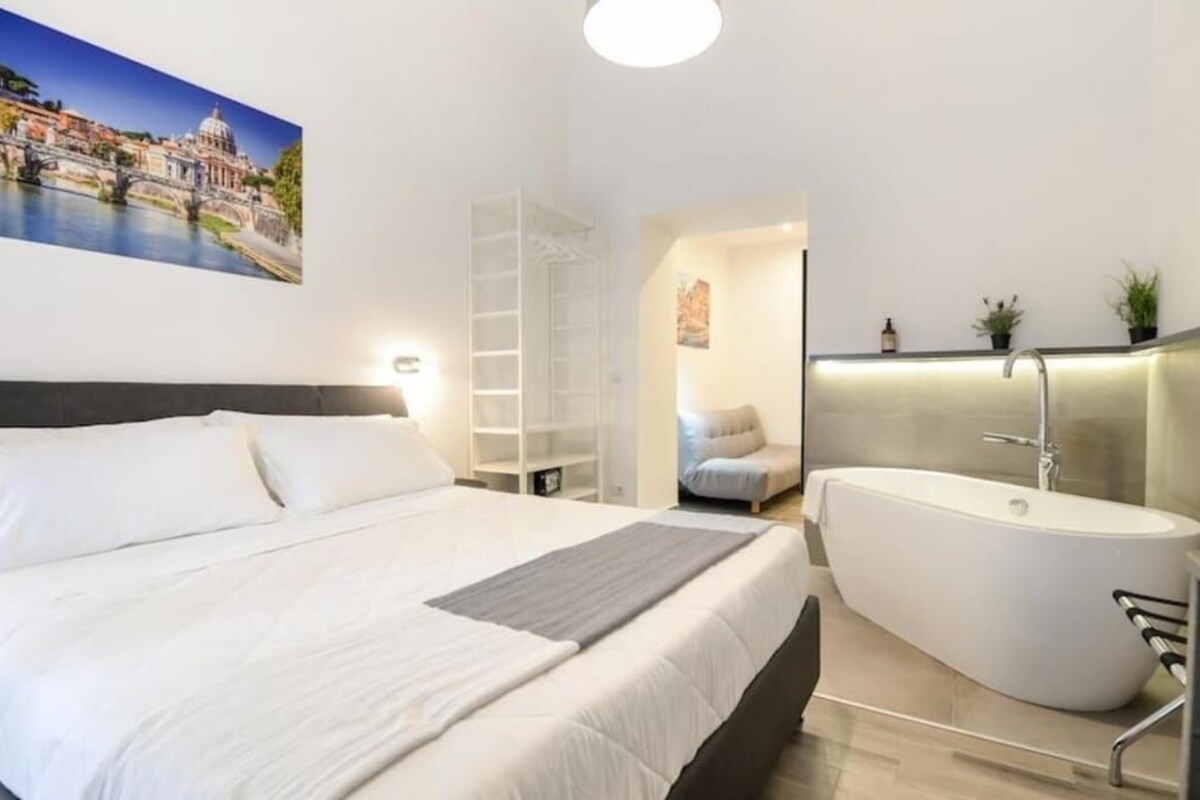 A modern bedroom with a large bed, a walk-in closet, a bathroom with a freestanding bathtub, and a cityscape painting on the wall.