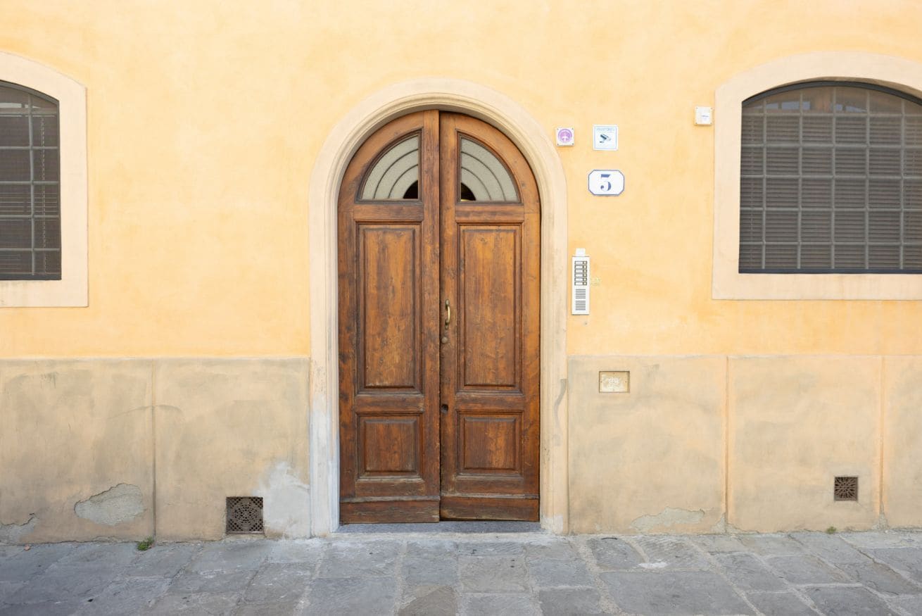 A double wooden door with a small window above it, set in a building with a plain facade and a stone-paved ground.