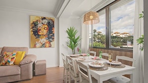 A dining area with a white table set for four, surrounded by white chairs, a large window with a view of buildings, and a painting on the wall. - Apartamento 'Brisa del Sur' para 6 personas, Huelva y Portugal, Wifi, A/C, luminoso (Ayamonte)