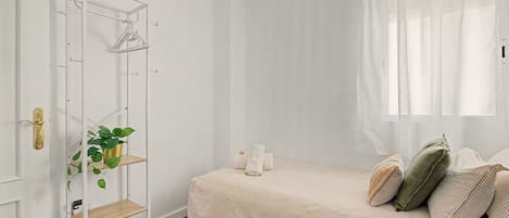 A bedroom with a bed, a white wardrobe, a plant shelf, and a window with sheer curtains.