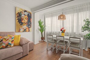 A living room with a sofa, a dining table with chairs, a large painting, and a potted plant. - Apartment 'Brisa del Sur' (Isla Canela) for 6 guests, Huelva & Portugal, Wi-Fi, A/C and bright (Ayamonte)