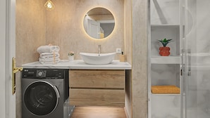 A compact bathroom with a built-in washing machine, a round mirror, and a wooden vanity. - Apartamento 'Brisa del Sur' para 6 personas, Huelva y Portugal, Wifi, A/C, luminoso (Ayamonte)