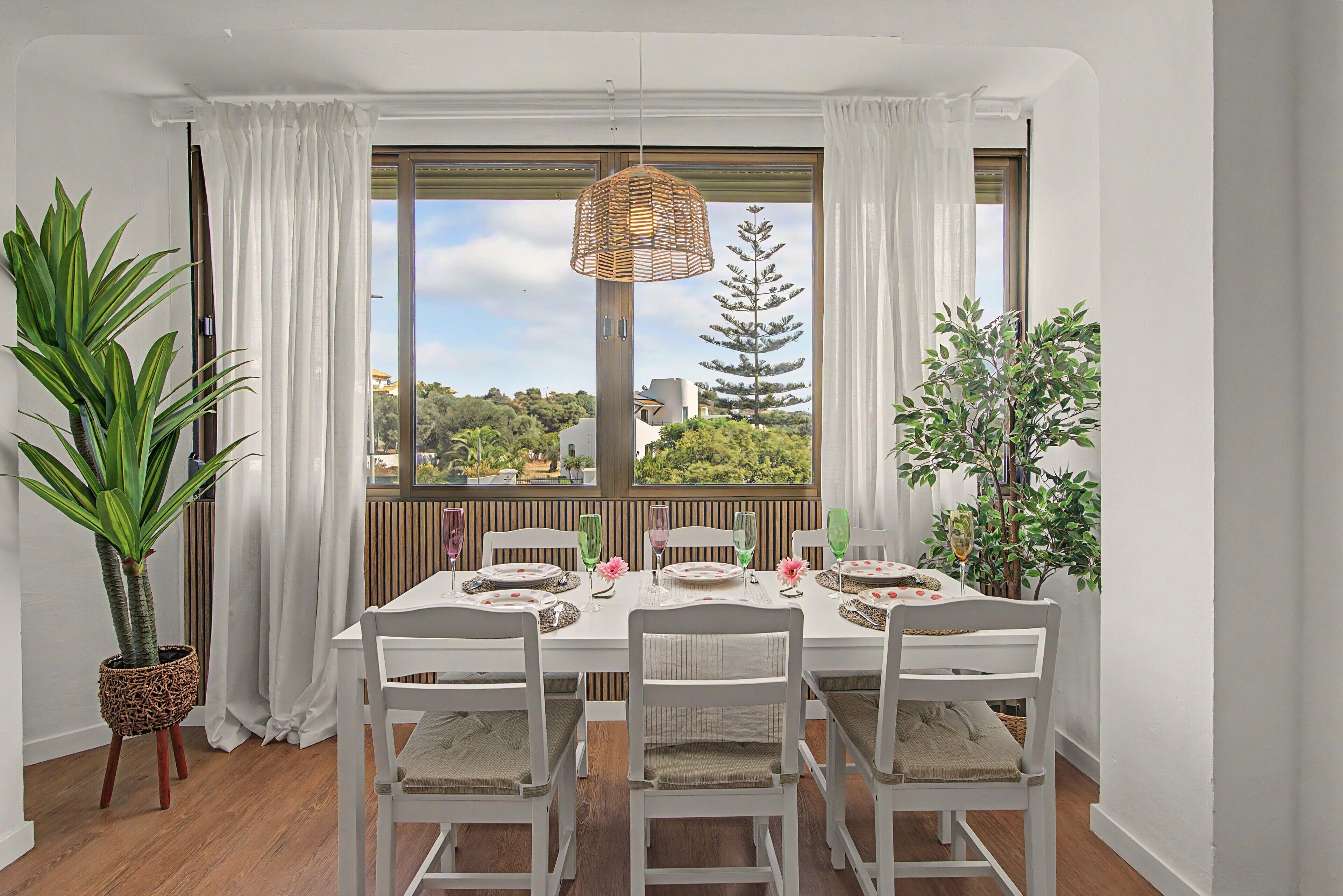 A dining area with a table set for four, white chairs, and a view of greenery through large windows.