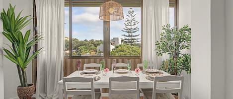 A dining area with a table set for four, white chairs, and a view of greenery through large windows.