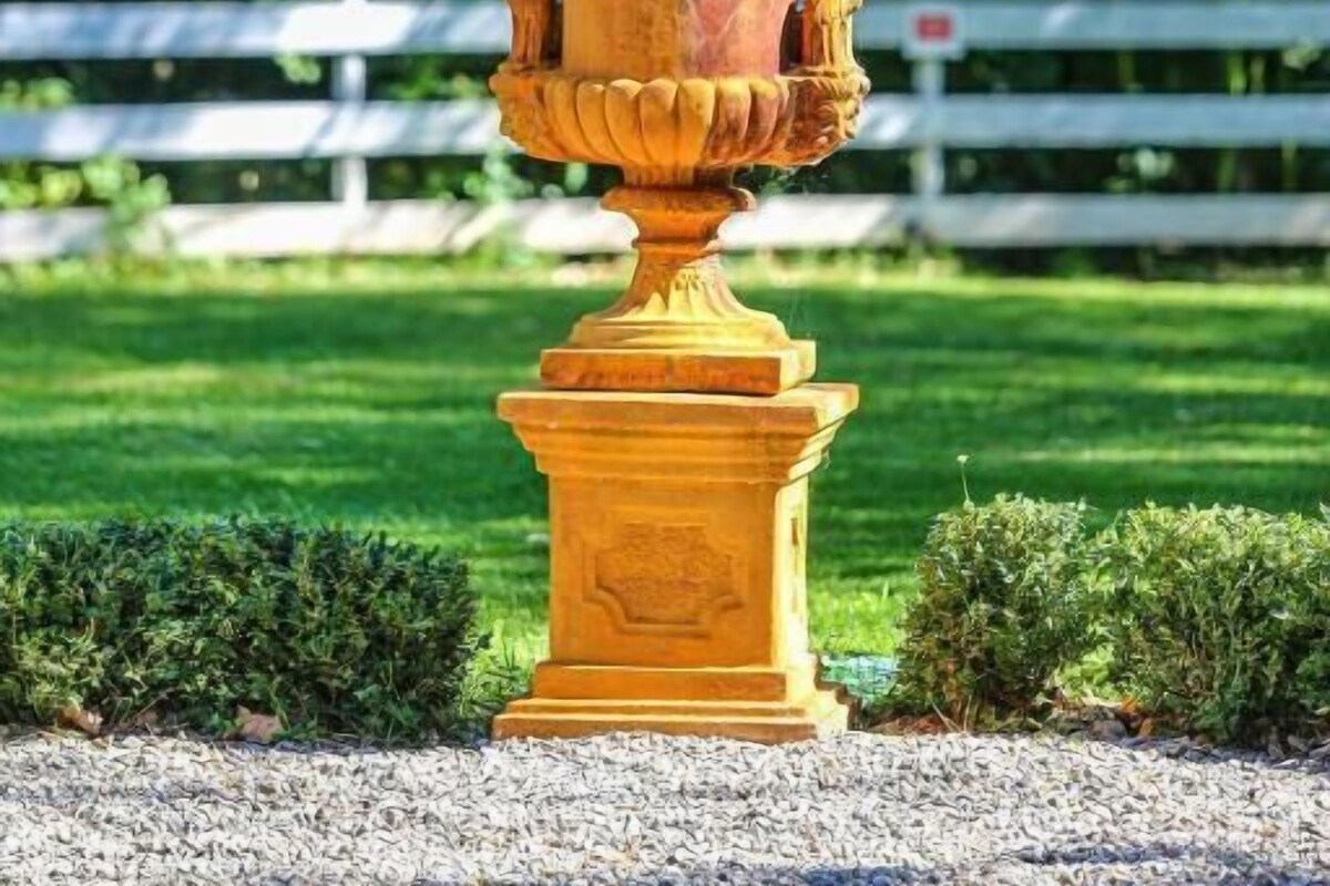 A yellow urn with a plant, set on a pedestal, surrounded by a gravel path and a white fence in a garden.