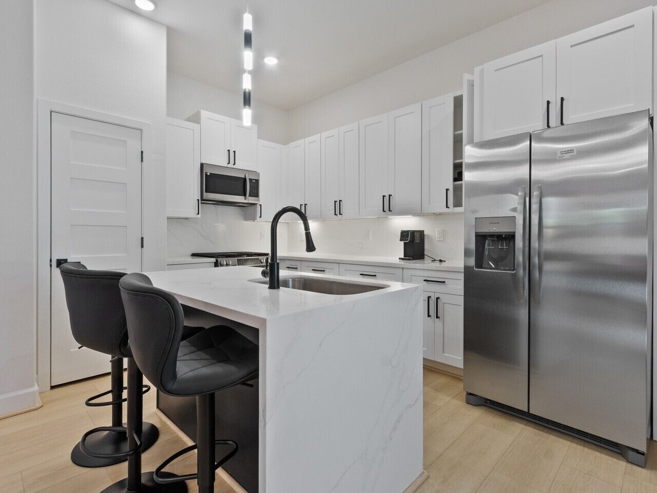 A modern kitchen with white cabinets, a stainless steel refrigerator, a built-in oven, and a marble island with bar stools.