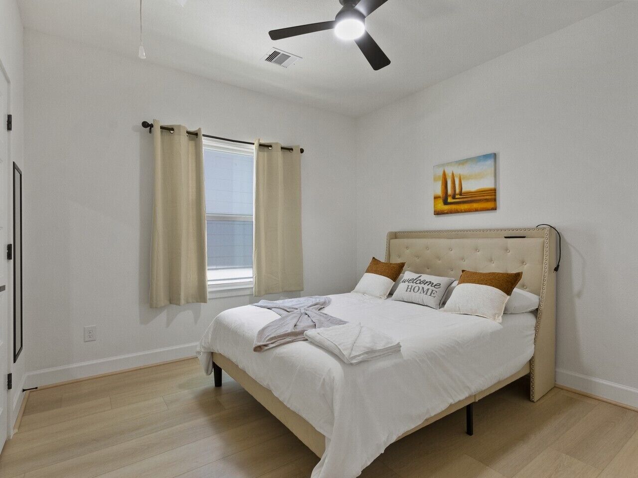 A bedroom with a bed, curtains, a ceiling fan, a painting, and a window.