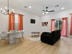 A modern living room with a dining area, a black sofa, a TV mounted on the wall, and orange curtains. - Oma Cozy Homes - A Modern, Newly Built Retreat in Houston (Houston)