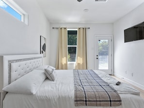 A bedroom with a bed, a window with curtains, a door, and a mounted TV. - Oma Cozy Homes (Houston)