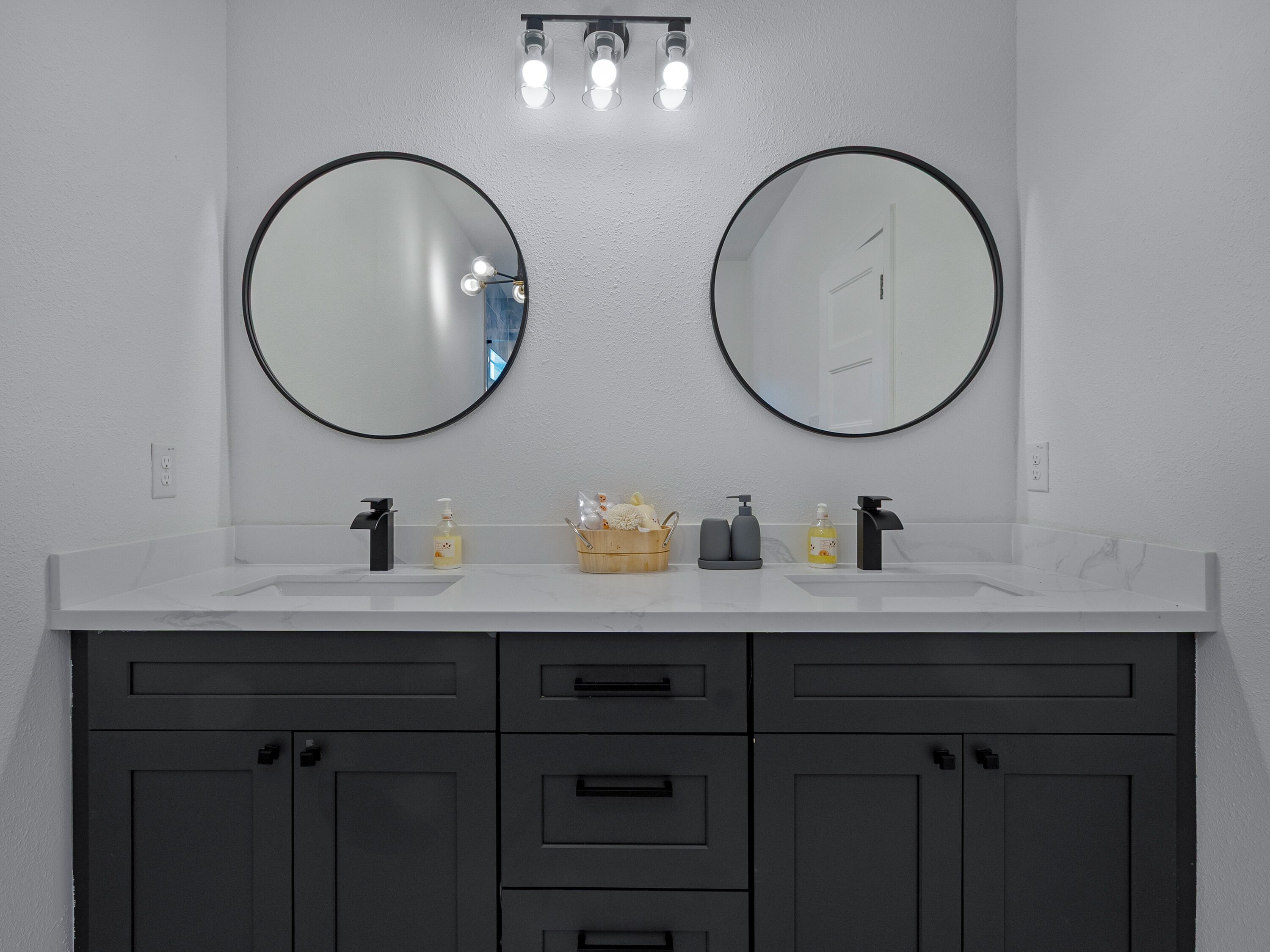 A bathroom with two round mirrors, a white countertop, and dark gray cabinets with drawers.