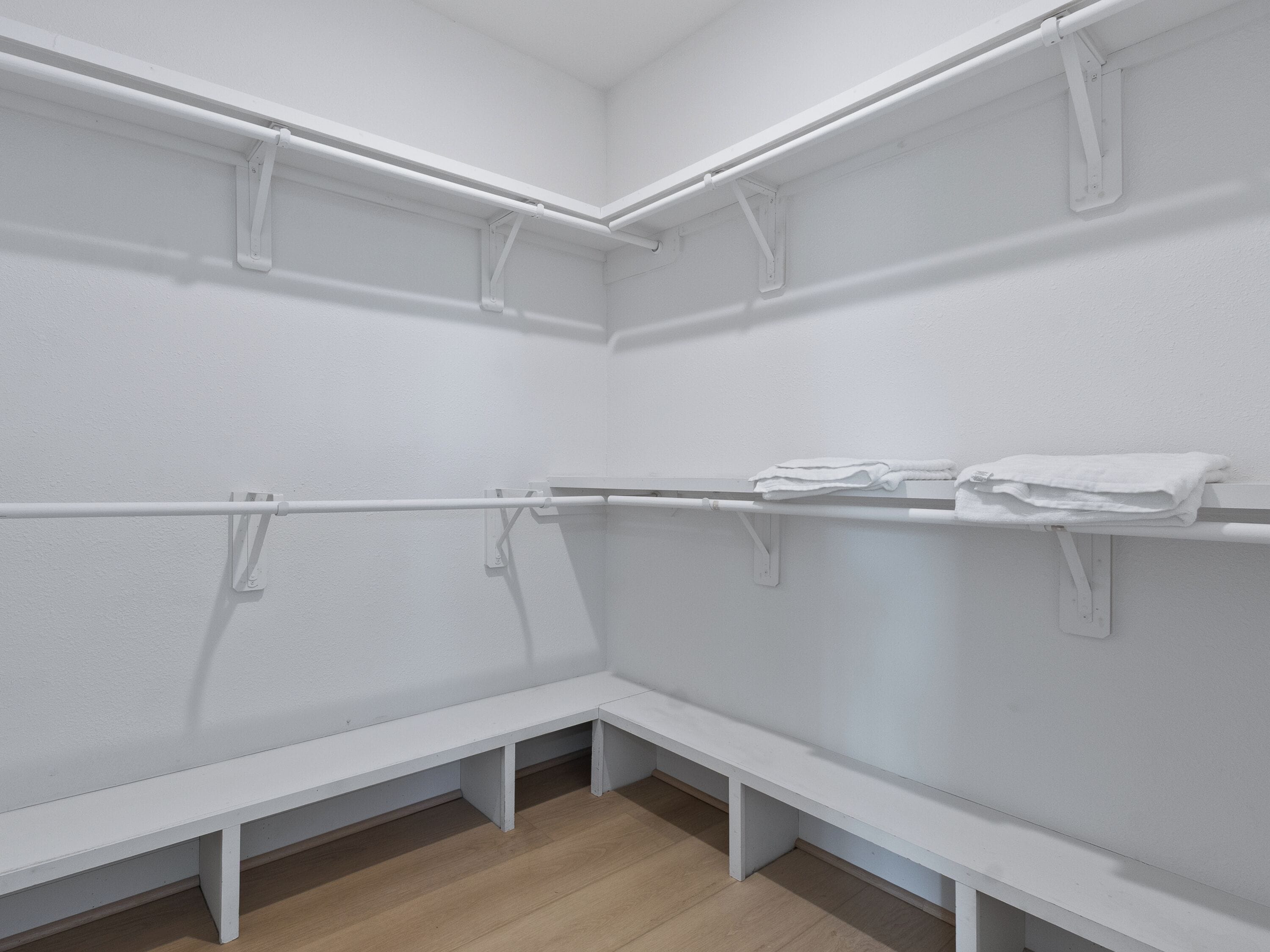 A white room with wooden flooring, white benches, and white shelves with towels.