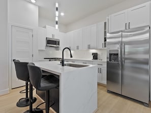 A modern kitchen with white cabinets, a stainless steel refrigerator, a built-in microwave, and a bar stool area. - Oma Cozy Homes (Houston)